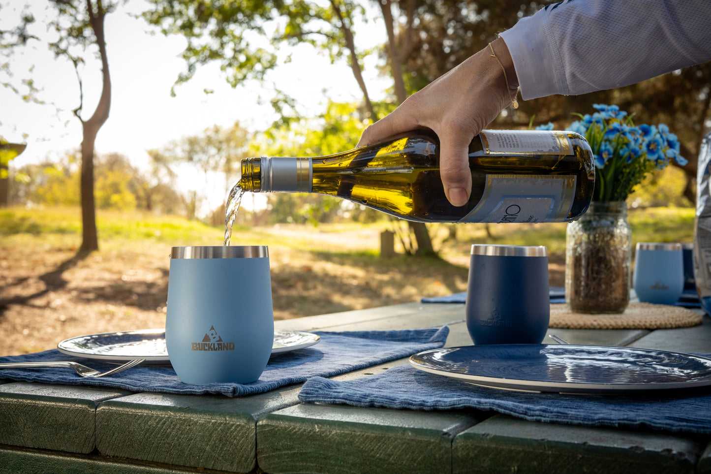Person pouring wine into blue 12 ounce Buckland tumblers on a picnic table outdoors.