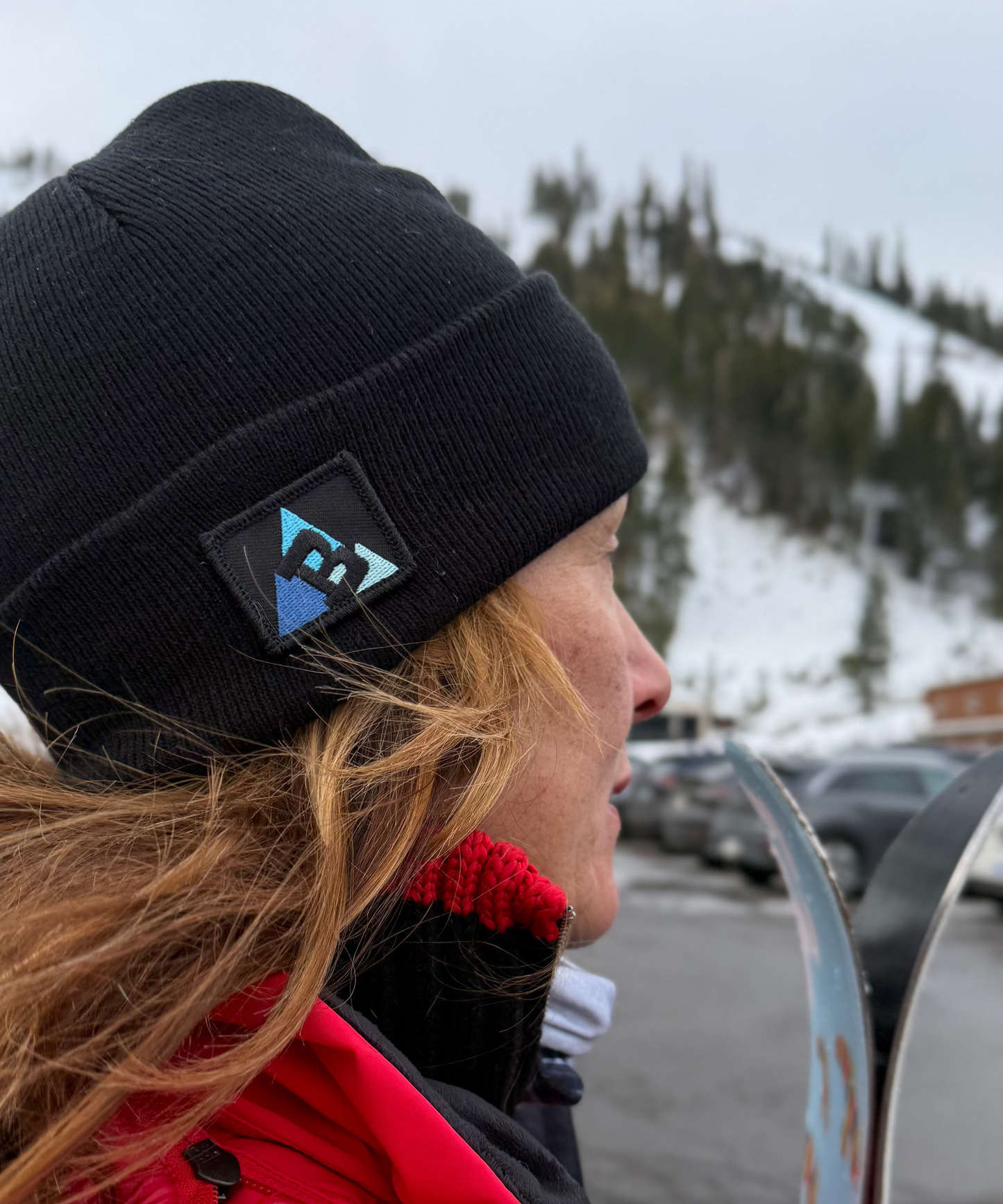 Person wearing a black beanie with a logo and red coat, standing outdoors with snowy landscape in the background.