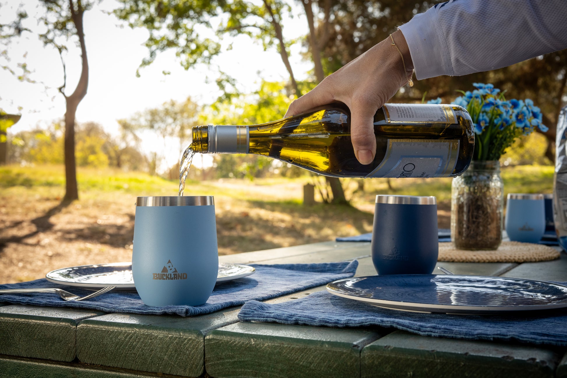 Person pouring wine into blue 12 ounce Buckland tumblers on a picnic table outdoors.