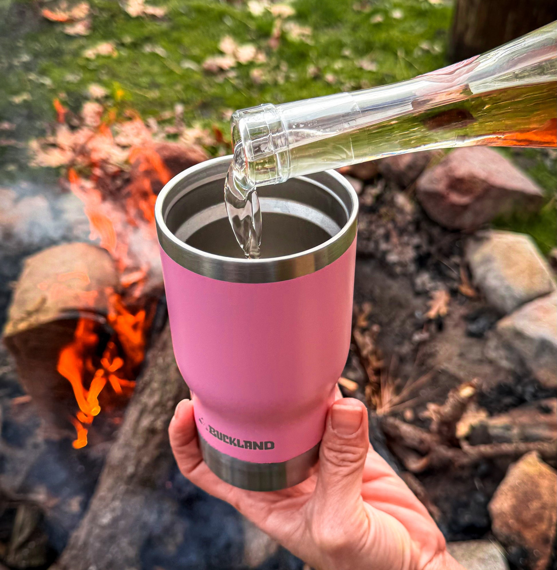 Pink insulated tumbler being filled with a clear liquid by a person outdoors near a campfire.