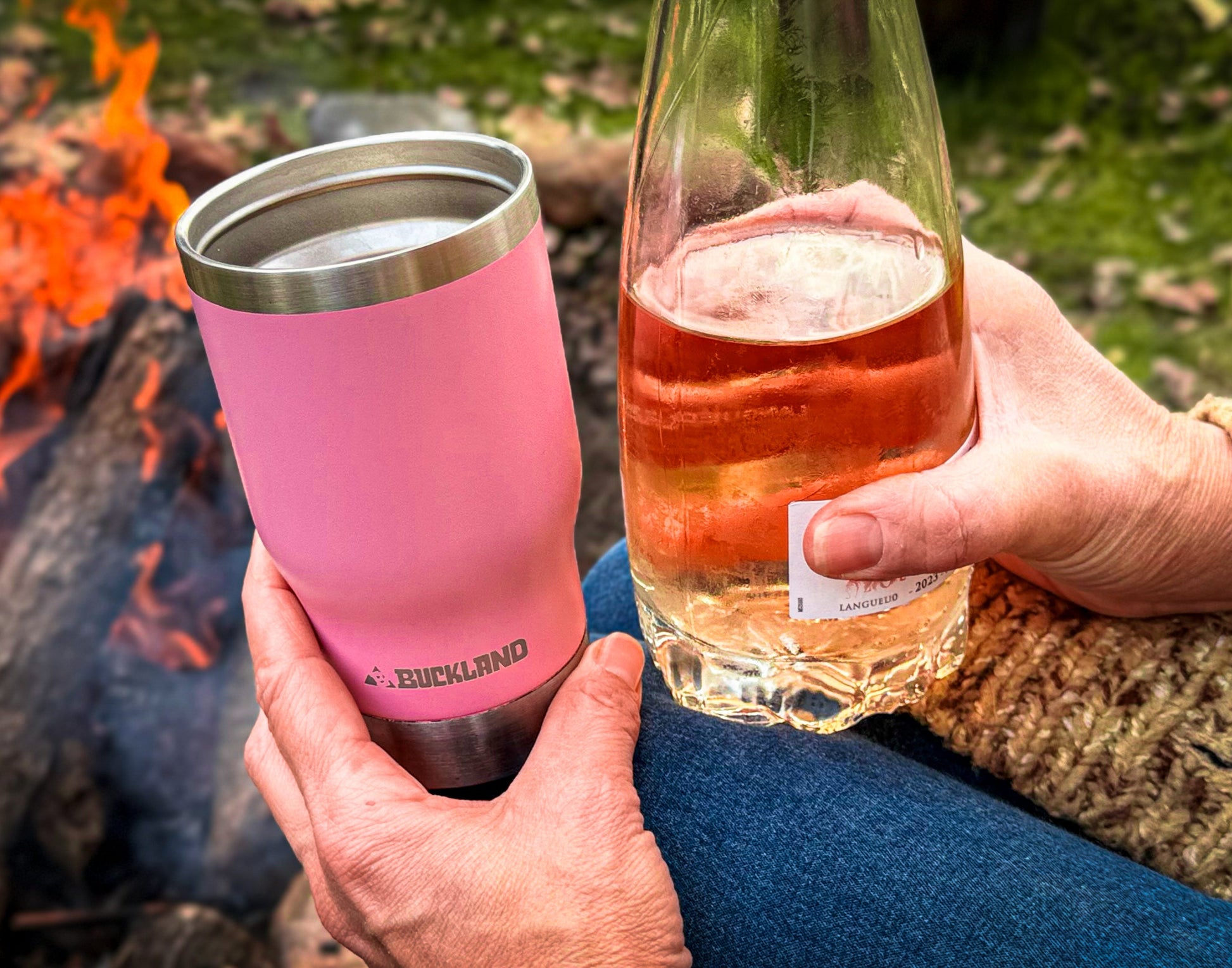 Person holding a pink insulated tumbler and a bottle of rosé by a campfire.